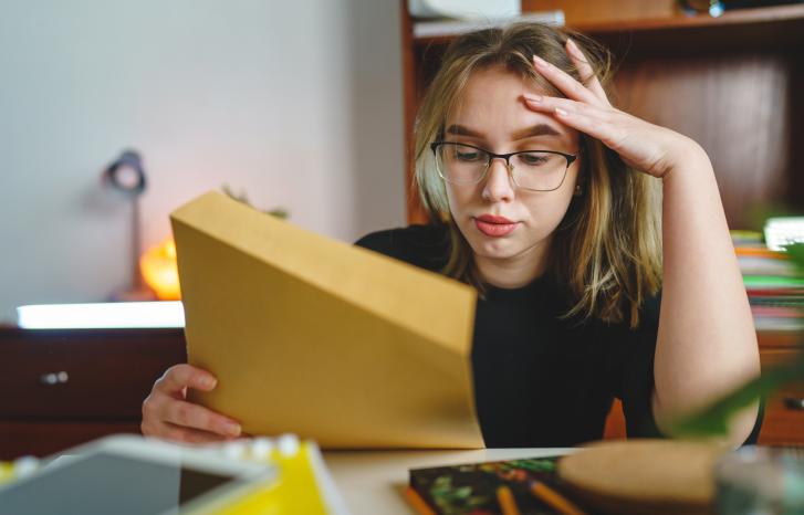 Jente eller ung kvinne med blondt og hår og briller som ser stresset ut og holder papir i den ene hånda