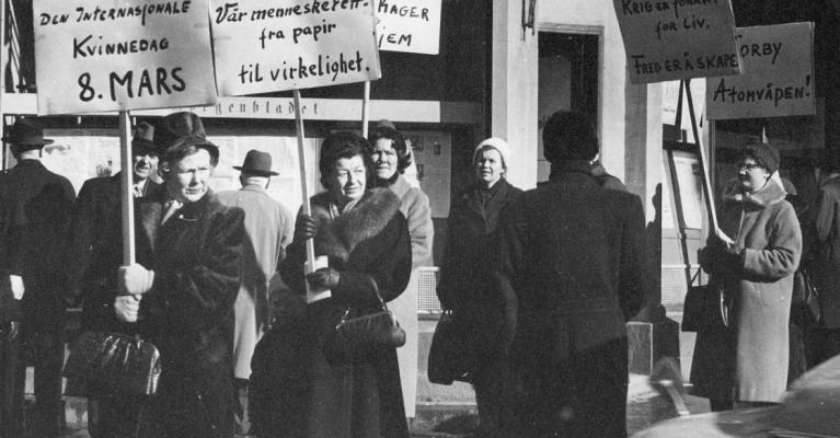 Åtte kvinner går langs fortauet med plakater på det som trolig er Norges første 8. mars markering. Parolene handler om likelønn, fred og nei til atomvåpen