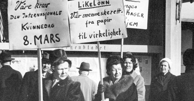 Svart-hvitt bilde av to kvinner i mørke kåper. De holder hver sin plakat med teksten "Våre krav den internasjonale kvinnedag 8. mars", og "Likelønn, vår menneskerett -fra papir til virkelighet".