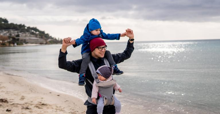 Pappa på tur på stranden med ett barn i bæresele og ett på nakken