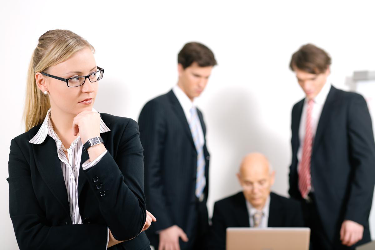 Woman in suit stands with her back to in front of three men talking in front of a computer without looking at her