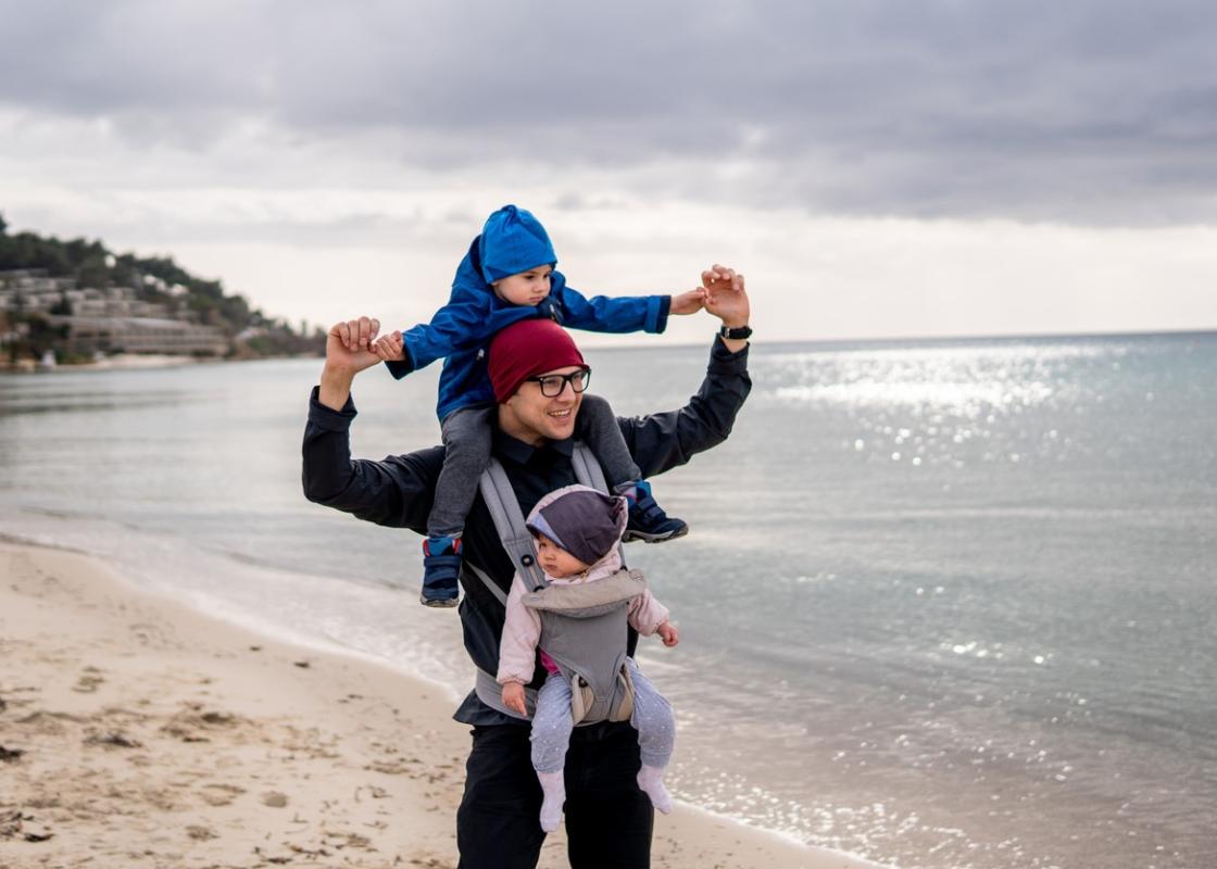 Pappa på tur på stranden med ett barn i bæresele og ett på nakken